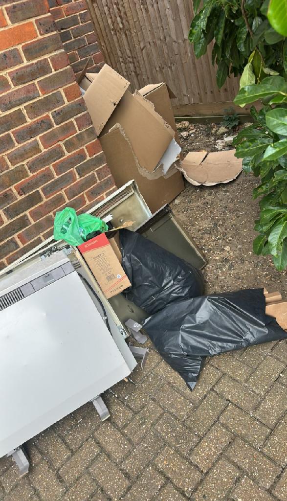 Before clearance in Bromley Town: an enclosed outdoor yard beside a brick wall and timber fencing with scattered cardboard pieces, general debris, and black bin liners on the paving near the corner.